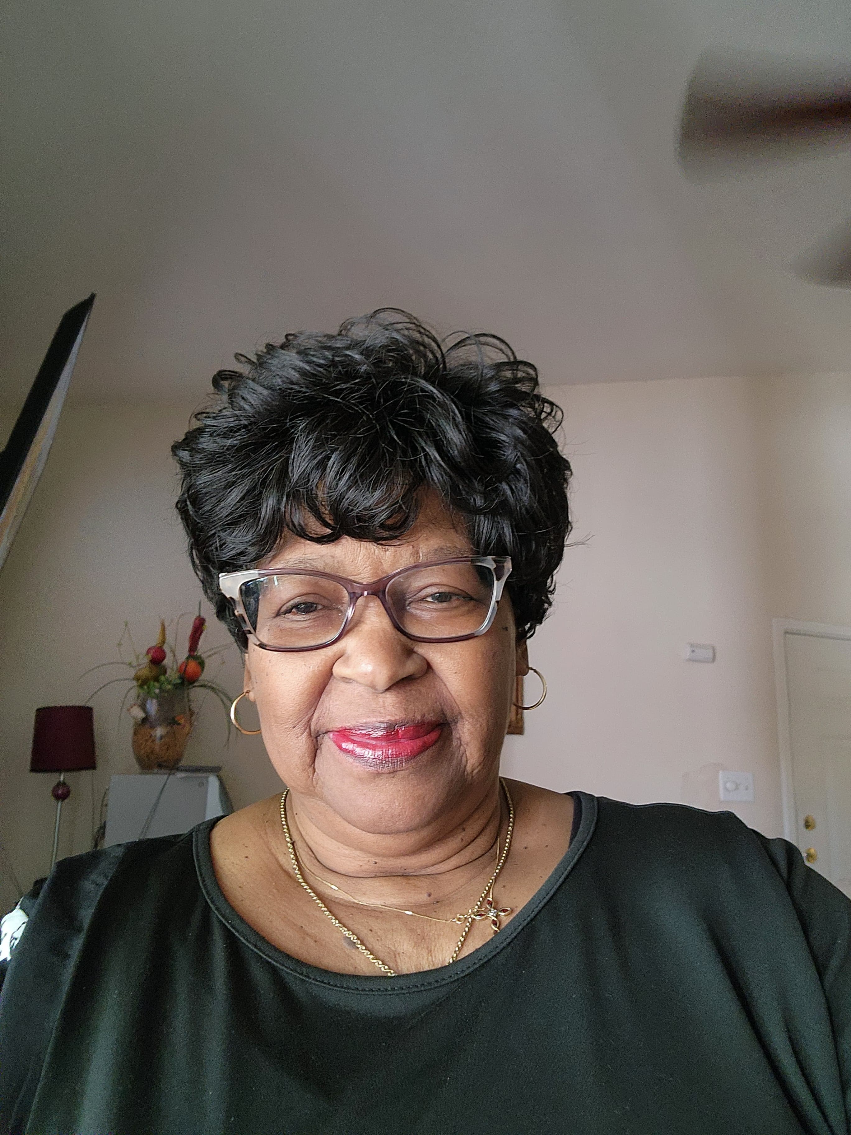 Smiling Black woman with short curly hair, glasses, red lipstick, and gold jewelry indoors.